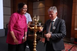 Two people stand next to a ceremonial lamp; a woman in a pink kurta and a man in a suit participate in a lamp-lighting ceremony at a formal event.