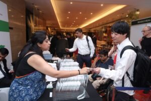 Conference registration desk scene, with a woman handing out badges to participants, including a man with a backpack and people in the background.