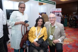 Three people pose together at an assistive technology exhibition booth, one seated in a wheelchair.