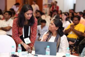Two women interacting at a conference table with a laptop, audience seated in the background.