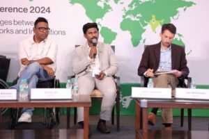 Three panelists seated on stage at the India Conference 2024, with map graphics in the background.