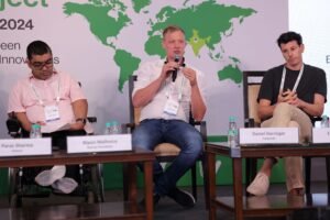 Three male panelists, one using a wheelchair, sit on stage at the India Conference 2024 with a world map in the background.